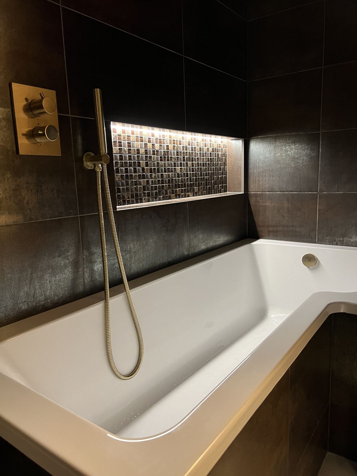 Dark bronze-tiled bathroom with an L-shaped shower bath, brushed brass shower and back-lit niche